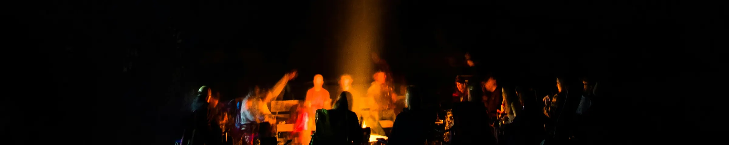 Menschen sitzen abends im Kreis um ein Lagerfeuer am Waldrand